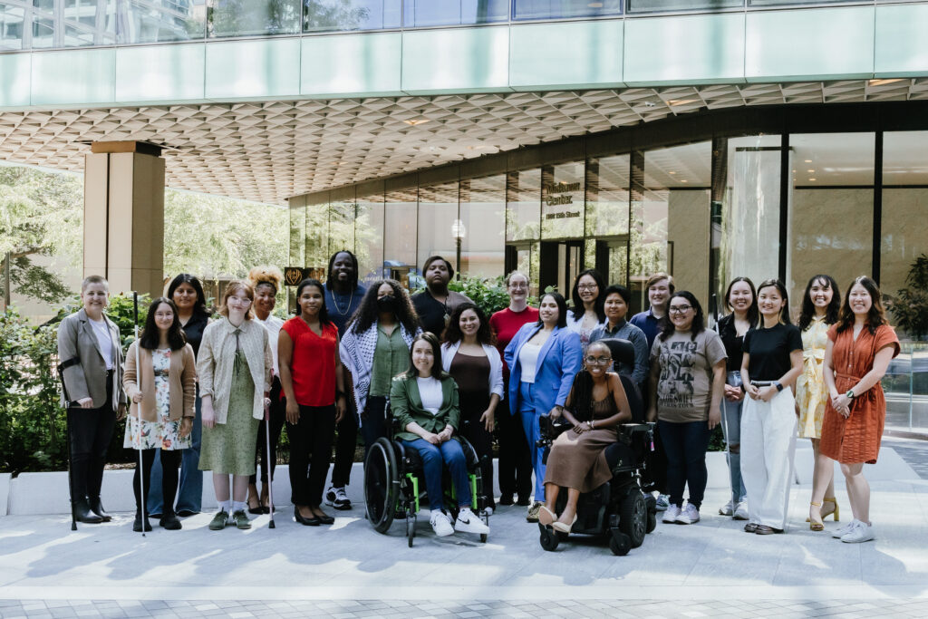 The 2024 AAPD Summer Interns and AAPD Programs Staff, a multiracial group of young people in their 20s stand in front of a metallic office building. They are wearing business casual clothing. Some of them are using a variety of mobility aids, including canes, crutches, and wheelchairs.