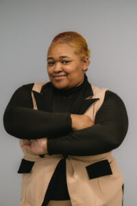 Ela’jah, a Black woman with short cropped brownish-red hair is standing in front of a gray wall and smiling with her mouth closed, with her arms crossed in front of her. She is wearing a black shirt and a tan professional blazer vest 