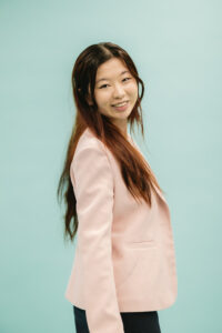 Janna, an Asian woman with long brown hair with auburn tones, is smiling and standing, turning to look over her shoulder toward the camera. She is wearing a pale pink blazer and a dark navy skirt. She is standing in front of a mint green background. 