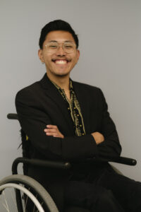 Huá Huī, a Chinese person in their 20s with black hair, is sitting in their wheelchair with their arms crossed and has a big smile on their face. They are wearing a black suit with a button-down shirt with yellow flowers on it. They have gold, geometric shaped glasses, and small gold-colored earrings. 