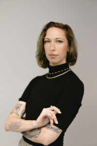 Lazy, a white person with green hair is posing in front of a light grey background with a serious, powerful expression and their arms. They have a nose septum piercing and are wearing a black short-sleeved turtleneck and two layered necklaces. Several of their arm tattoos are visible.