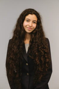 Brianna, a Latina woman in her 20s with long, dark brown hair is standing in front of a grey background, wearing a black blazer and grey dress. Her head is slightly tilted to the side, and she is smiling.