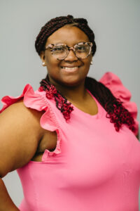 Lauryn Huffman, a Black woman with braids with red curled ends and tortiseshell glasses is wearing a pink top with ruffle sleeves and smiling at the camera in front of a grey background. 