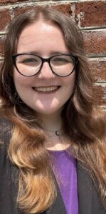 A selfie of Adriana, a white woman in her 20s with long brown wavy hair and black glasses, is smiling at the camera. She is wearing a purple shirt and black blazer, and is in front of a brick wall