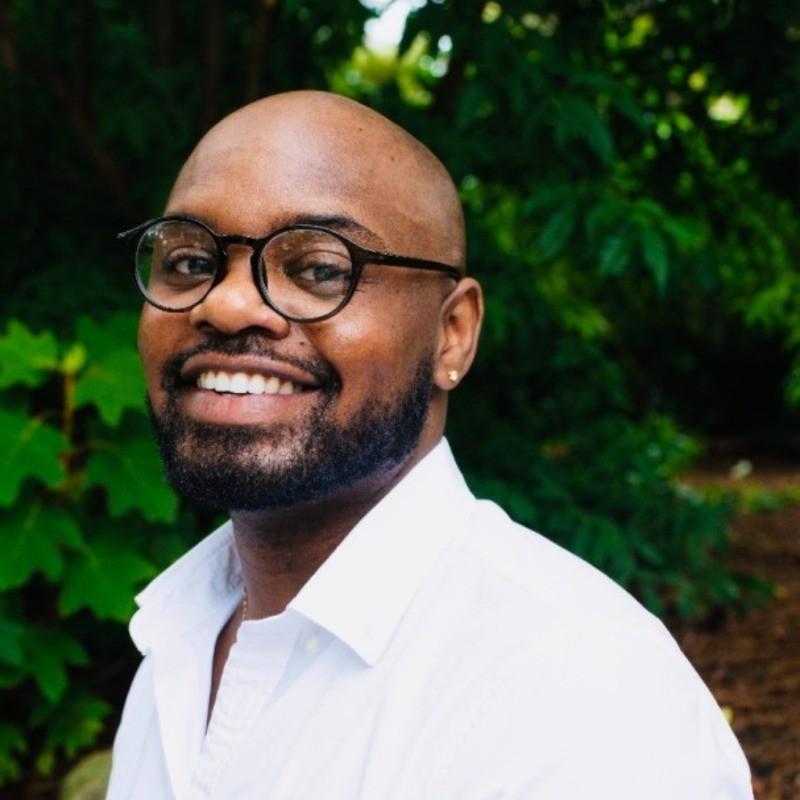 A Black man with a bald head, neatly trimmed beard, and mustache is smiling warmly at the camera. He is wearing round black eyeglasses and a crisp white button-down shirt, open at the collar. A small stud earring is visible in his left ear. His teeth are showing in a broad smile, giving the photo a friendly and approachable feel. He is photographed outdoors in daylight, surrounded by lush greenery with vibrant leaves and trees that create a natural backdrop. The lighting is soft and even, highlighting his face clearly against the deep green background.