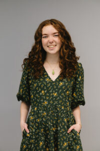 Dylan, a white femme person in their 20s with medium length curly red hair and freckles is smiling at the camera with her hands in her pockets in front of a solid gray background. They are wearing a dark green v-neck dress that is dotted with small orange flowers.