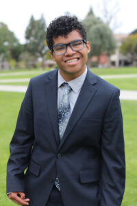 Full body photo of Jason Green in a navy blue suit, grey shirt and tie with a mixture of blues and grey. Background is of a grass area on California State University Long Beach's campus