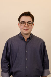 ID: Maddox is a white man with short brown hair and rose-tinted glasses wearing a dark blue button down shirt and smiling.