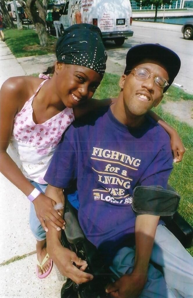 Two Black people smiling at the camera. One is a wheelchair user, the other is leaning against him.