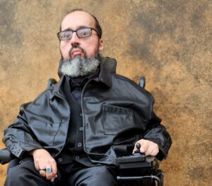 Headshot of Valois Vera. Valois is a person sitting in their power chair, with a black leather jacket, black suit vest, and a black button up, along with black pants. They are also wearing rings. He has short brown hair, black rectangular glasses, and a brown and white mustache and beard.